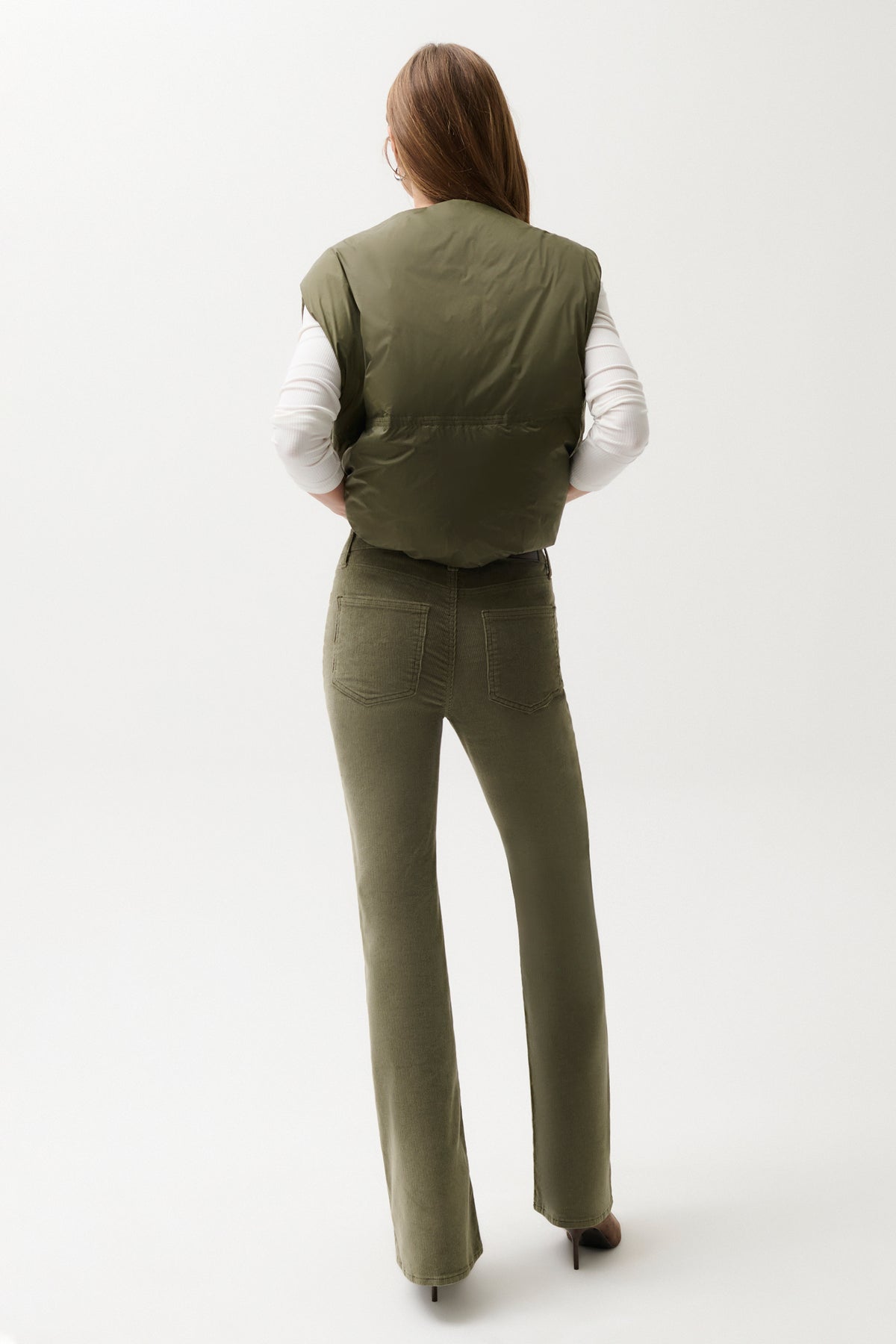 A woman shown from the back wearing an olive green puffer vest over a white long-sleeve top and matching olive green pants, photographed against a clean white studio background.